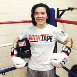 This is an image of Dr. Khanita posing in a boxing ring in a white t-shirt and grey shorts. The t-shirt says “RockTape: Go stronger, longer” She is wearing white padded boxing gloves and is holding a black padded boxing helmet with the word Title across it. It appears as though she just finished sparring.