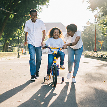 This is an image of a dad and mom helping their daughter learn to ride on a bike with training wheels.